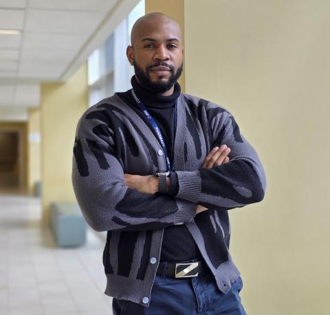 Man with a beard wearing a grey and black sweater is standing confidently in a hallway with his arms crossed
