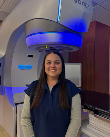 A woman standing in front of a machine. She has brown hair, wearing a blue scrub shirt over a grey long sleeve