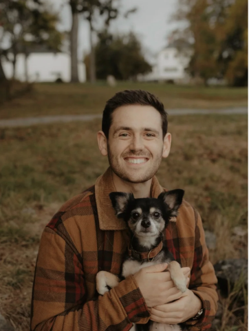 Man with brown hair holding a black and brown dog. The man is wearing a brown plaid jacket. 