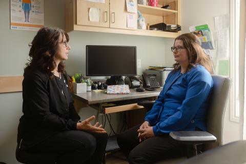 two women in a office woman on left is wearing all black, woman on right wearing a blue long sleeve with black pants