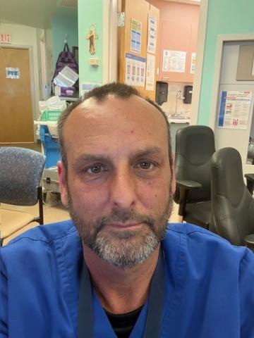 Male with brown short hair and grey beard. He is wearing blue scrubs.