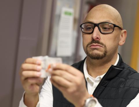 Man with glasses holding a label. He is wearing a black vest over a white long sleeve shirt